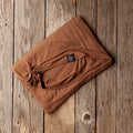 A folded brown fabric item, likely a blanket or throw, displayed diagonally on a rustic wooden plank background. A small black brand tag is visible on the fabric.