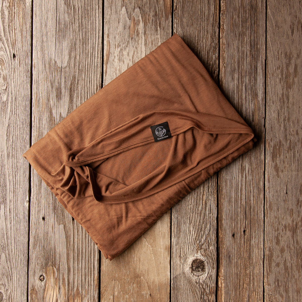 A folded brown fabric item, likely a blanket or throw, displayed diagonally on a rustic wooden plank background. A small black brand tag is visible on the fabric.