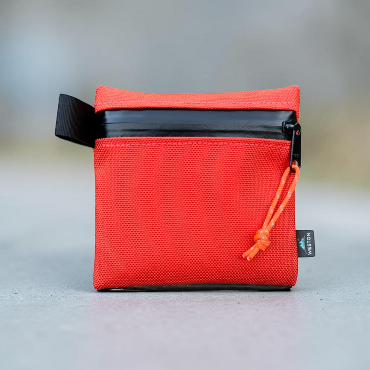 A small red textured pouch with a black zipper, black webbing loop, and an orange drawstring, photographed on a smooth gray surface with a blurred background.
