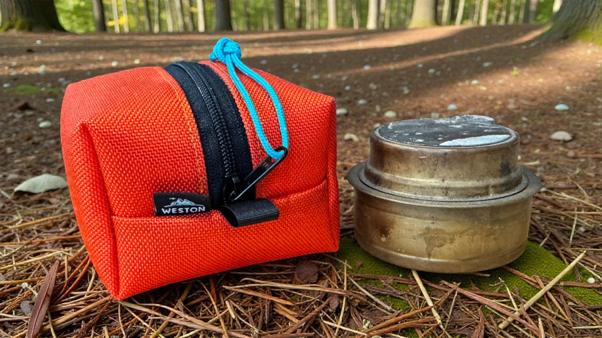 Orange pouch with a blue drawstring and black zipper, featuring the Weston brand logo, next to a metal container on a forest floor.