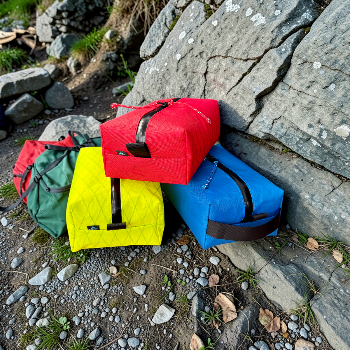 Three quilted dry bags in red, yellow, and blue, along with a green bag in the background, arranged on the ground against a stone wall outdoors.
