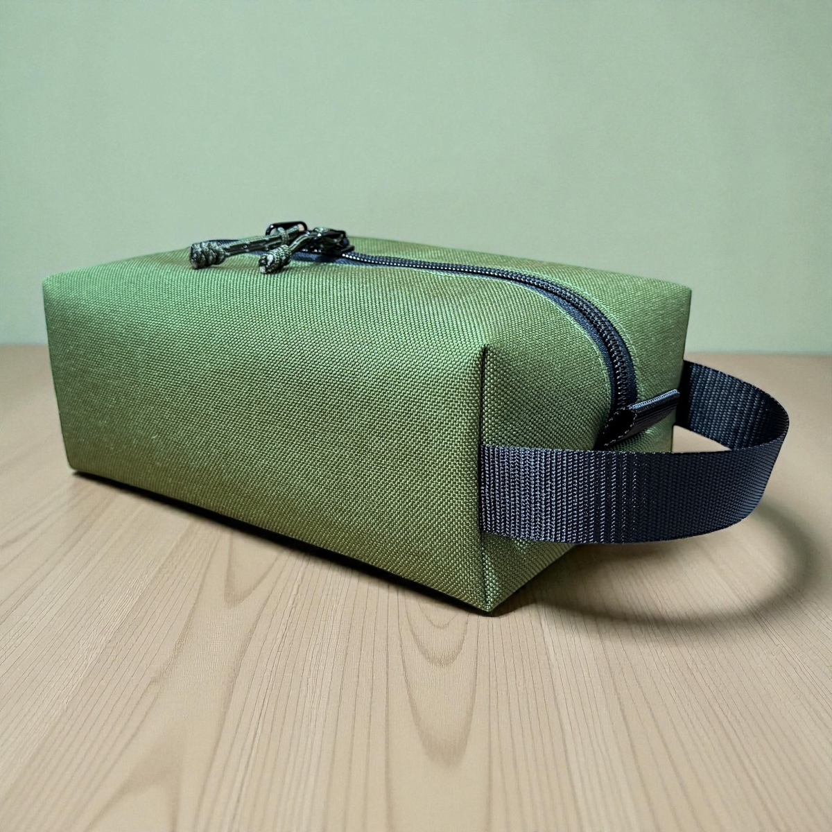 Green toiletry bag with a black strap on a wooden surface and light green background