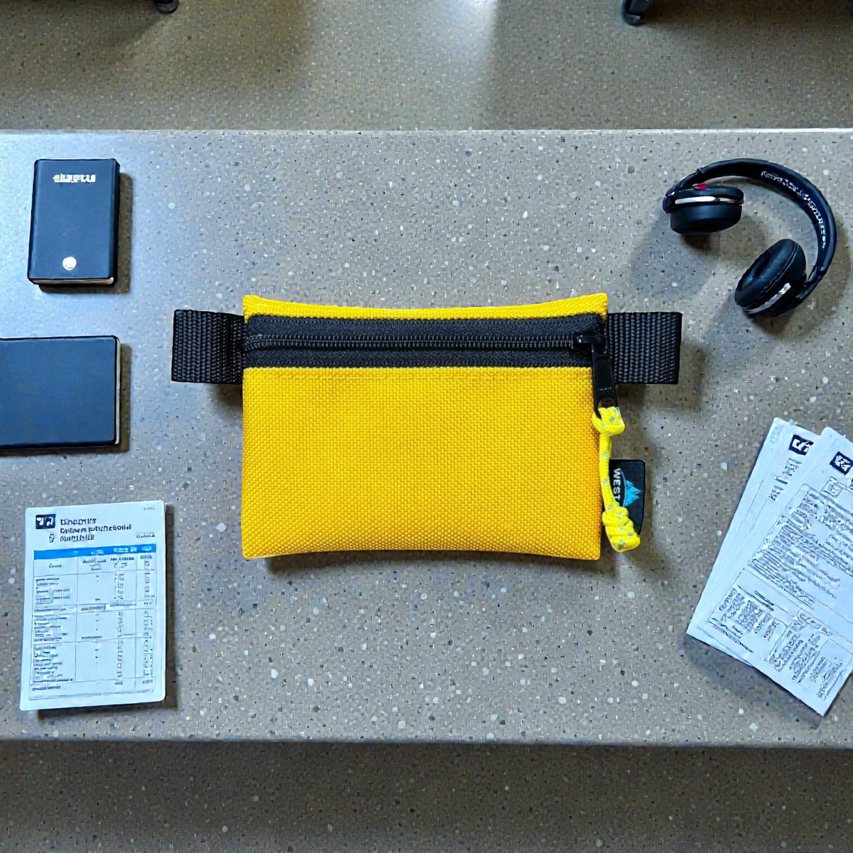 A bright yellow mesh zippered pouch with black webbing straps and a black zipper, laid flat on a speckled gray countertop. Surrounding it are various travel items including a USB drive, headphones, passport photos, and boarding passes.