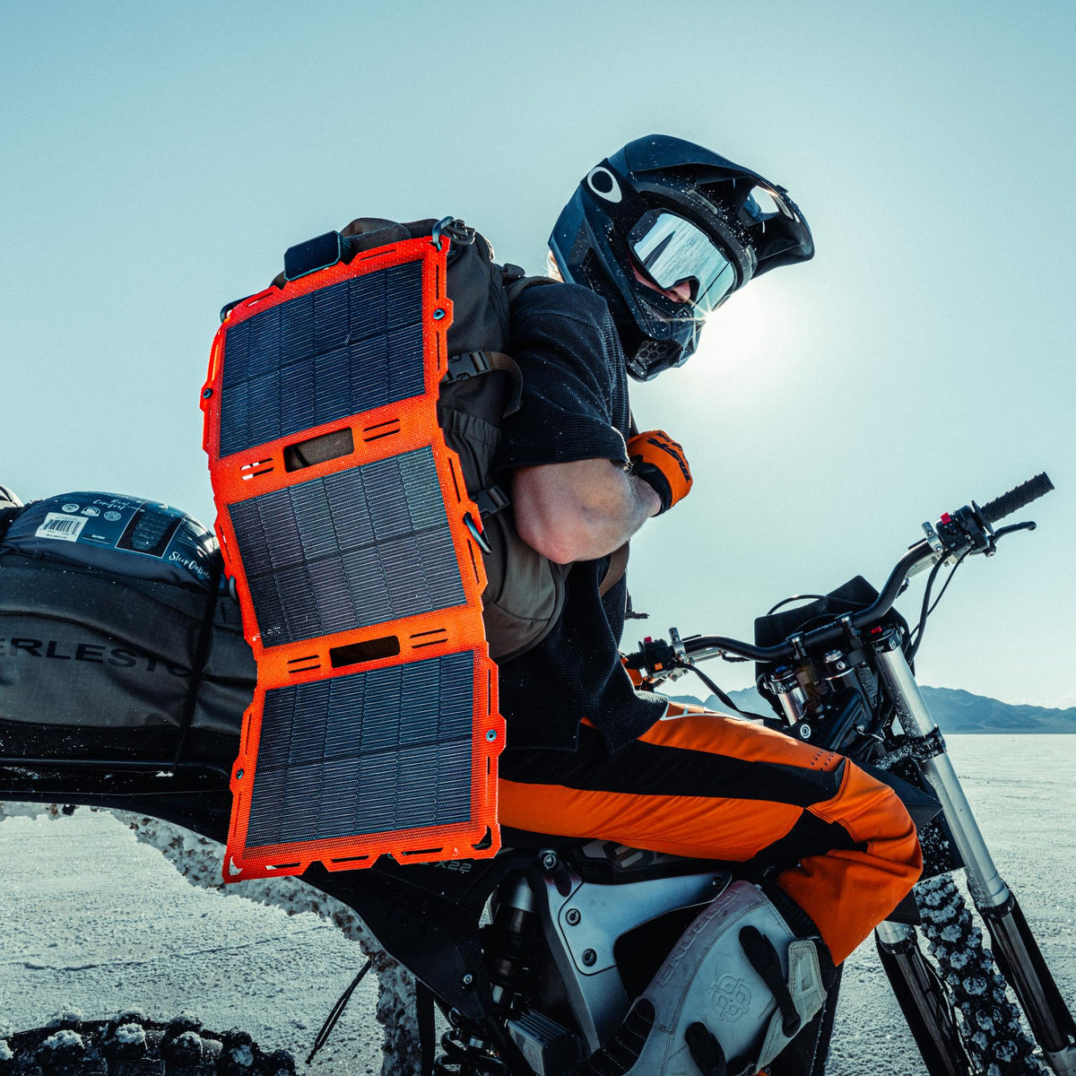 A person wearing a black helmet and goggles sits on a motorcycle in a bright outdoor setting, carrying a large foldable backpack solar panel charger on their back. The charger has three orange-framed panels with dark blue solar cells.