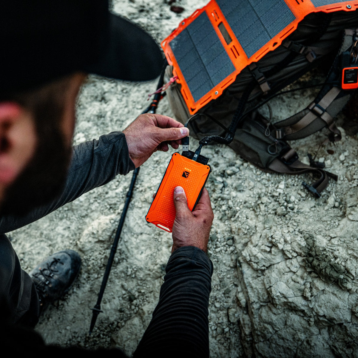 Person using an orange portable device to charge a solar panel on a rocky surface