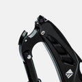 Close-up of a black metal climbing or mountaineering accessory with cutout panels and a small white geometric logo near the bottom right, photographed on a light gray background.