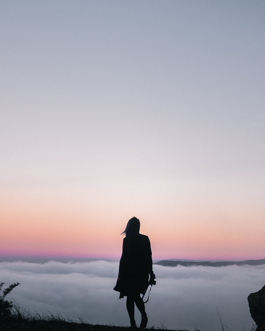A woman stands in silhouette on a cliffside overlooking a sea of clouds at sunset, with a soft pink and purple sky above.