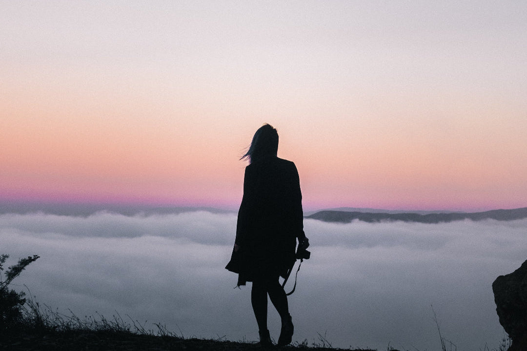 A woman stands in silhouette on a cliffside overlooking a sea of clouds at sunset, with a soft pink and purple sky above.